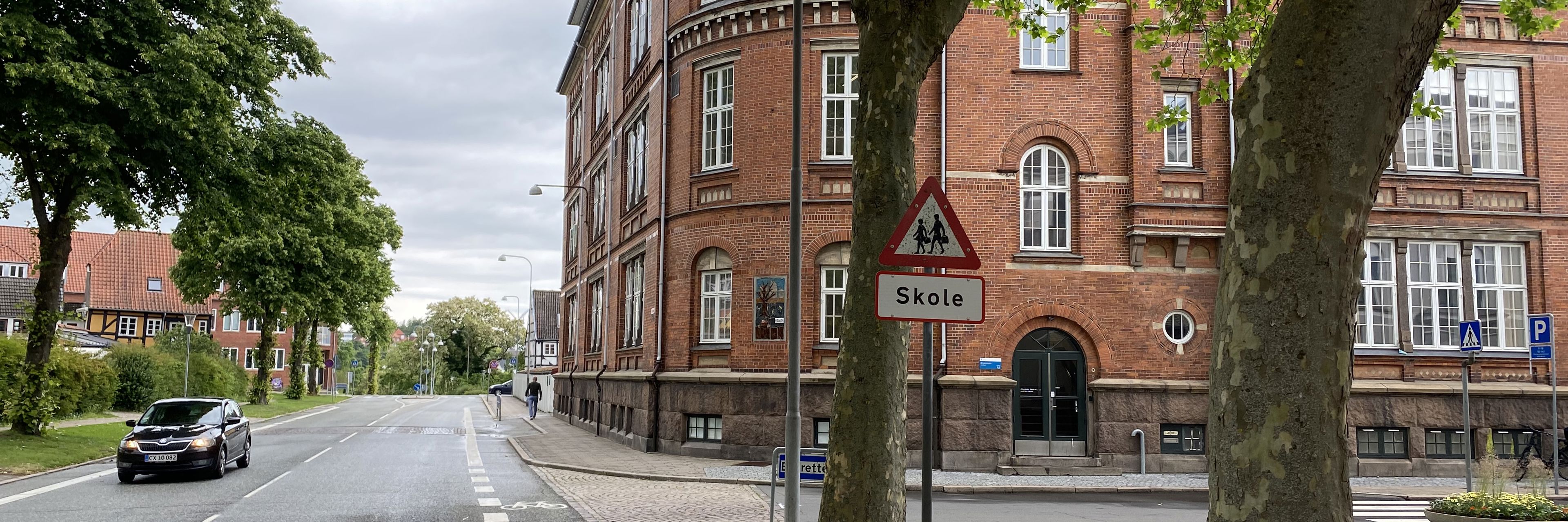 Ørkildskolen afdeling Byen - en gammel rød murstensbygning set fra vejen - udsynet er delvis blokeret med træer i forgrunden og det klassiske trekantede skilt for skole