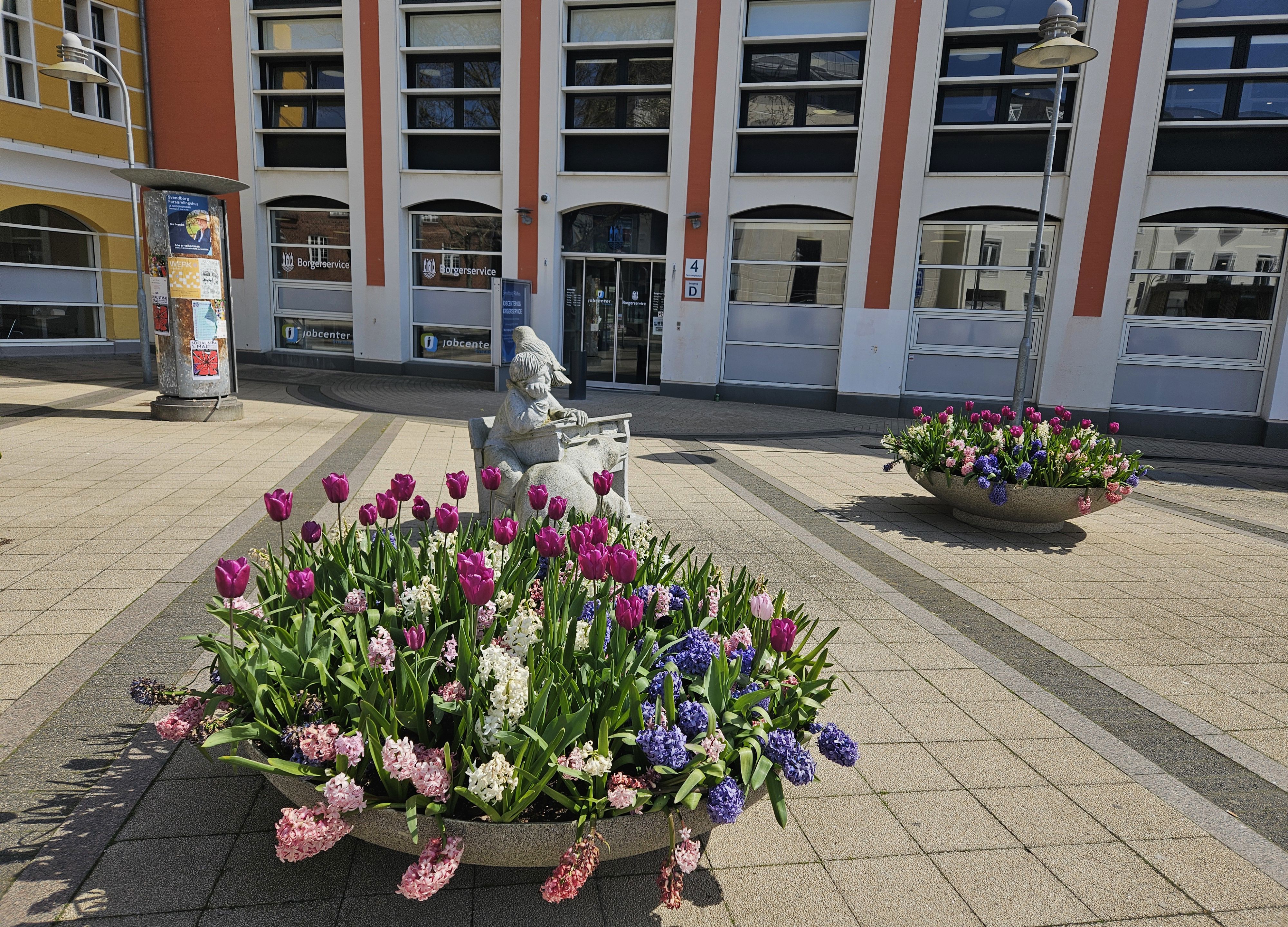 En kumme med forårsblomster foran indgangen til en stor bygning, der er hvid og rød.